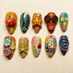 May include: A collection of ten uniquely designed artificial nails. Each nail features a different design, including a red apple, a starfish, and a ladybug. Colors include yellow, blue, orange, and pink. Some nails have button and star embellishments.