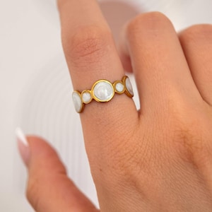 Puede incluir: Un anillo de tono dorado que presenta una serie de cinco engastes circulares de tamaños variables, cada uno incrustado con un material blanco nacarado y liso. La banda está construida con un marco de metal continuo que sujeta los elementos.