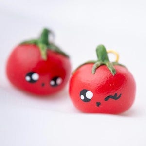 Peut inclure: Deux breloques rouges en forme de tomate avec des tiges vertes et des visages de dessins animés mignons. Une breloque fait un clin d'œil. Ces breloques sont probablement des boucles d'oreilles ou des pendentifs.