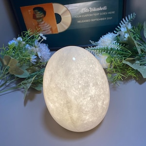 Könnte beinhalten: Eine leuchtende, eiförmige dekorative Lampe mit einer strukturierten, weißen Oberfläche. Die Lampe ist der Mittelpunkt, vor dem Hintergrund eines gerahmten Albums und floralen Akzenten. Das Albumcover ist orange und der Text auf dem Rahmen lautet "YOUR CUSTOM TEXT GOES HERE RELEASED SEPTEMBER 2021".