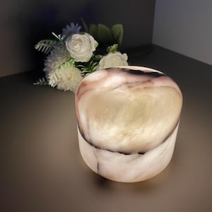 Puede incluir: Un portavelas cilíndrico de piedra translúcida con la parte superior redondeada. La piedra tiene una mezcla de vetas blancas, beige y marrón oscuro. Un ramo de flores blancas se encuentra en el fondo. El portavelas mide unos 10 cm de alto.