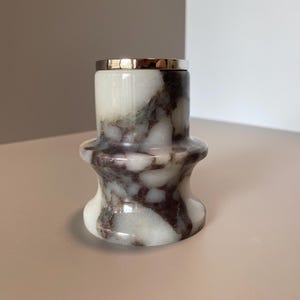 Könnte beinhalten: Ein dekorativer Kerzenhalter aus Marmor mit silbernem Rand. Der Kerzenhalter ist weiß mit dunkelbraunen und grauen Adern. Das Design ist zylindrisch mit ausgestelltem Boden und Oberteil.