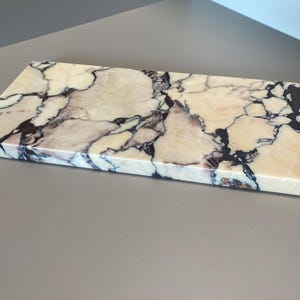 Peut inclure: Planche de service rectangulaire en marbre avec une base crème et des veines complexes violettes et noires. La planche a une finition polie, adaptée au service des aliments. Dimensions : environ 30 cm x 20 cm.