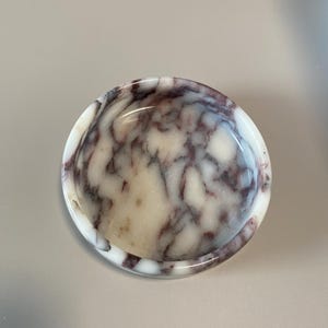Könnte beinhalten: Eine runde Marmorschale mit einem weißen und burgunderfarbenen Marmormuster. Die Schale hat ein flaches, geschwungenes Design und eignet sich zum Halten kleiner Gegenstände. Die Schale besteht aus poliertem Stein.