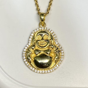 Könnte beinhalten: Eine goldfarbene Halskette mit Anhänger, die eine lächelnde Buddha-Figur zeigt. Der Anhänger ist mit kleinen, klaren Edelsteinen umrandet. Die Halskette ist ebenfalls goldfarben, was ein stimmiges und elegantes Schmuckstück ergibt.