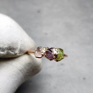 May include: A rose gold ring featuring three raw gemstones in pink, purple, and green. The gemstones are set in a textured gold setting, creating a unique and natural look. The ring is a delicate piece of jewelry.