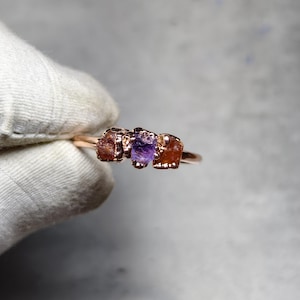 May include: A rose gold ring with three raw gemstones: two reddish-brown and one purple. The gemstones are set in a delicate, open-work design. The ring is shown against a neutral background.