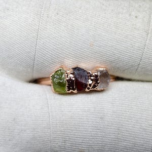 May include: A rose gold ring featuring three raw gemstones: a green peridot, a deep red garnet, and a clear quartz. The ring's band and settings have a textured, organic design, showcasing the natural beauty of the stones.