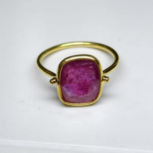 May include: A gold ring with a large, square-cut, deep pink gemstone. The ring has a simple band and a bezel setting that highlights the stone's colour and shape. The gemstone is a vibrant, eye-catching focal point.