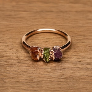 May include: A rose gold ring featuring three small, raw, uncut gemstones set in a row. The stones are an orange-brown citrine, a light green peridot, and a purple amethyst, each held in place by a textured metal setting on a thin, smooth band.
