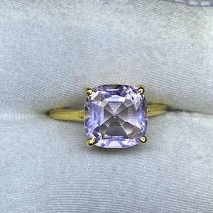 May include: A gold ring featuring a large, square-cut, light purple gemstone. The gemstone is set with four gold prongs. The ring band is a simple, polished gold.