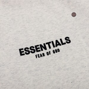 May include: Close-up of a light gray fabric with the black text "ESSENTIALS" and "FEAR OF GOD" printed diagonally across the bottom. The fabric has a subtle texture.