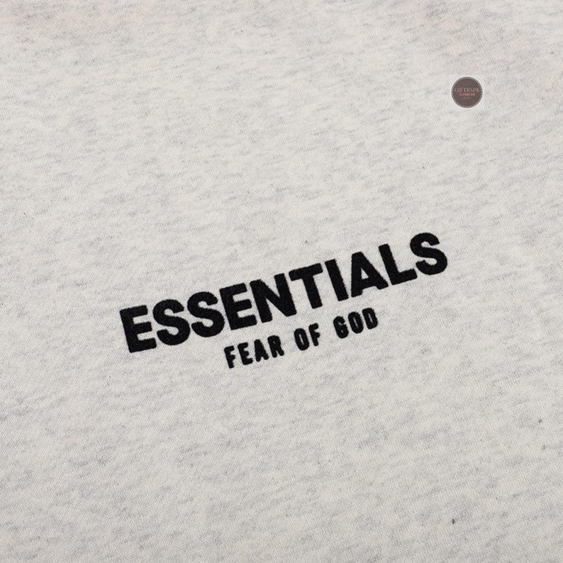 May include: Close-up of a light gray fabric with the black text "ESSENTIALS" and "FEAR OF GOD" printed diagonally across the bottom. The fabric has a subtle texture.