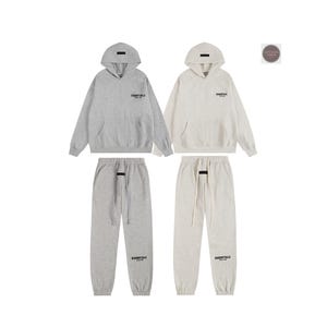 May include: Two matching gray and cream-colored hoodie and sweatpants sets. Each hoodie has a hood and a front pocket. The sweatpants have a drawstring and the word "ESSENTIALS" printed on the front.