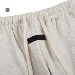 K&ouml;nnte beinhalten: Nahaufnahme einer hellgrauen Jogginghose mit Kordelzug und einem schwarzen Etikett mit der Aufschrift "ESSENTIALS". Die Hose hat eine lockere Passform und ist aus einem weichen, bequemen Material gefertigt. Der Kordelzug ist cremefarben.