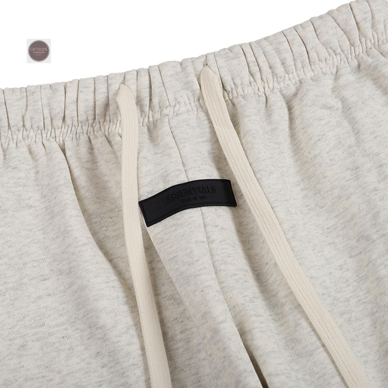 K&ouml;nnte beinhalten: Nahaufnahme einer hellgrauen Jogginghose mit Kordelzug und einem schwarzen Etikett mit der Aufschrift "ESSENTIALS". Die Hose hat eine lockere Passform und ist aus einem weichen, bequemen Material gefertigt. Der Kordelzug ist cremefarben.