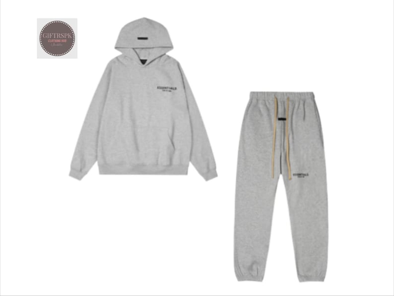 May include: A light gray hoodie and matching sweatpants set. The hoodie has a hood and the word "ESSENTIALS" printed on the chest. The sweatpants have a drawstring and the same logo. A casual, comfortable outfit.