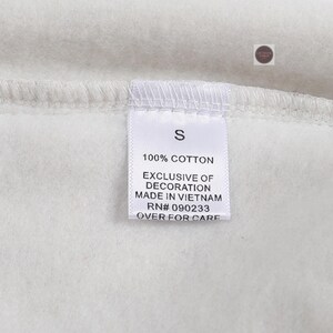 K&ouml;nnte beinhalten: Nahaufnahme eines wei&szlig;en Kleidungsst&uuml;cketiketts mit dem Buchstaben "S" aufgedruckt. Das Etikett enth&auml;lt auch den Text "100% COTTON", "EXCLUSIVE OF DECORATION", "MADE IN VIETNAM", "RN# 090233" und "OVER FOR CARE".