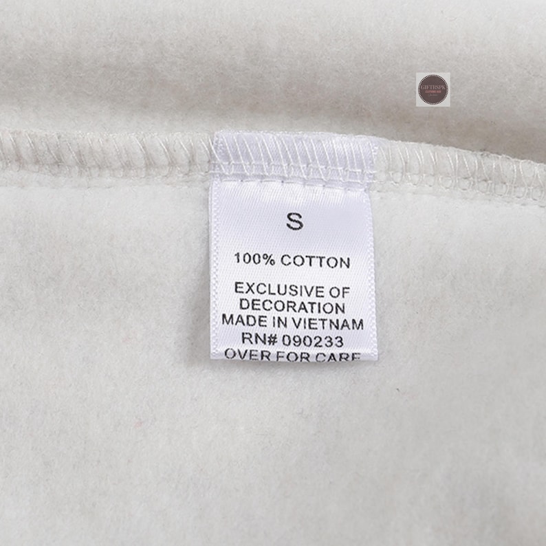 K&ouml;nnte beinhalten: Nahaufnahme eines wei&szlig;en Kleidungsst&uuml;cketiketts mit dem Buchstaben "S" aufgedruckt. Das Etikett enth&auml;lt auch den Text "100% COTTON", "EXCLUSIVE OF DECORATION", "MADE IN VIETNAM", "RN# 090233" und "OVER FOR CARE".