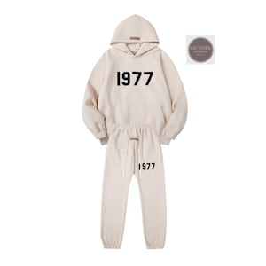 May include: A cream-colored hoodie and sweatpants set. The hoodie has a front pocket and a hood. The number "1977" is printed in black on the front of the hoodie and sweatpants. The sweatpants have a drawstring waist.