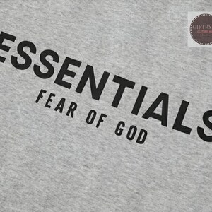 May include: A light gray fabric with the word "ESSENTIALS" printed in large, black capital letters, with "FEAR OF GOD" printed below. A small circular logo is in the upper right corner.