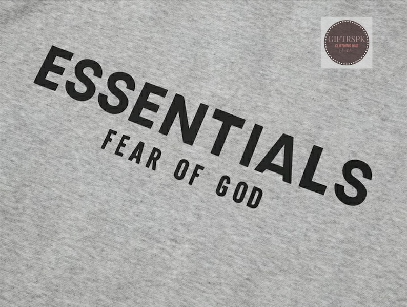 May include: A light gray fabric with the word "ESSENTIALS" printed in large, black capital letters, with "FEAR OF GOD" printed below. A small circular logo is in the upper right corner.