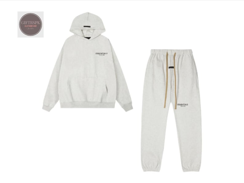 May include: A light grey hoodie and matching sweatpants set. The hoodie has a hood and a front pocket. The sweatpants have a drawstring waist. Both pieces have the word "ESSENTIALS" printed on them in black.