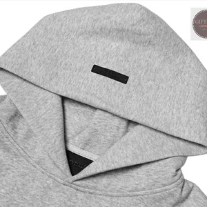 May include: Close-up of a gray hoodie with a hood. The hoodie has a black rectangular tag on the hood. The fabric appears to be a soft, textured material. The image is well-lit, highlighting the details of the garment.