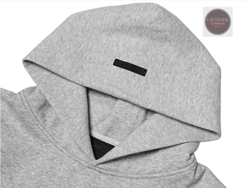 May include: Close-up of a gray hoodie with a hood. The hoodie has a black rectangular tag on the hood. The fabric appears to be a soft, textured material. The image is well-lit, highlighting the details of the garment.