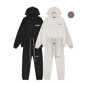 May include: Two matching tracksuits, one black and one light gray, each consisting of a hooded sweatshirt and sweatpants. The sweatshirts and sweatpants have the word "ESSENTIALS" printed on them. The sweatpants have drawstrings.