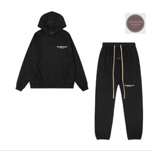 May include: Black hoodie and sweatpants set. The hoodie has a hood and a front pocket. The sweatpants have a drawstring waist. Both pieces have the word "ESSENTIALS" printed in white.
