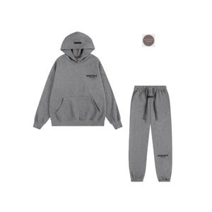 K&ouml;nnte beinhalten: Ein graues Hoodie- und Jogginghosen-Set. Der Hoodie hat eine Vordertasche und das Wort "ESSENTIALS" auf der Brust aufgedruckt. Die Jogginghose hat einen Kordelzug in der Taille und das Wort "ESSENTIALS" auf dem Bein aufgedruckt. Das Set ist aus weichem Material.