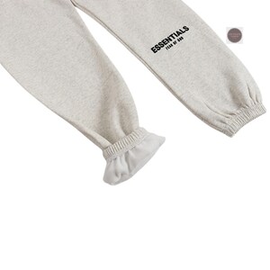 K&ouml;nnte beinhalten: Hellgraue Jogginghose mit elastischen B&uuml;ndchen und dem Wort "ESSENTIALS" in Schwarz auf dem Bein aufgedruckt. Die Hose ist aus einem weichen, bequemen Material gefertigt.
