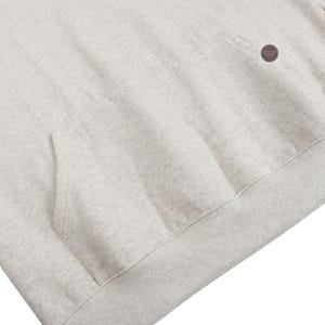 May include: Close-up of a light gray hoodie with a kangaroo pocket and a ribbed hem. The fabric has a speckled texture. A small, circular tag is visible near the shoulder.