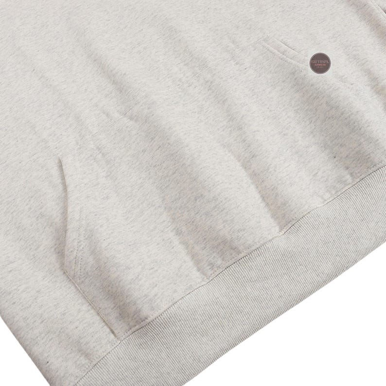 May include: Close-up of a light gray hoodie with a kangaroo pocket and a ribbed hem. The fabric has a speckled texture. A small, circular tag is visible near the shoulder.