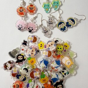 May include: A selection of colourful acrylic earrings featuring cartoon character designs. The earrings have silver-coloured hooks and come in various shapes, including animals and people. The designs include orange, blue, pink, and green characters.
