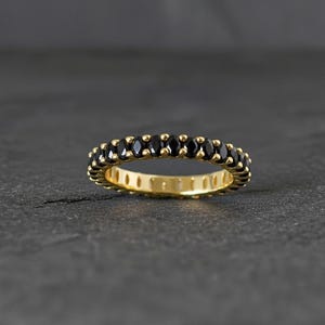 Puede incluir: Un anillo de oro con una banda de piedras negras. El anillo se encuentra sobre un fondo gris oscuro, resaltando el contraste entre el oro y el negro. Las piedras están espaciadas uniformemente.