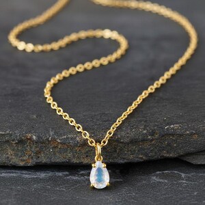 Peut inclure: Un collier doré délicat avec un petit pendentif en forme de poire. Le pendentif a un aspect iridescent bleu clair et blanc. La chaîne du collier est fine et repose sur une surface sombre et texturée.