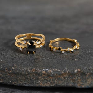 May include: Two gold rings. One ring features a large, black, teardrop-shaped stone with small, clear accent stones. The other ring is a thin band with small black and clear stones. Both rings are on a dark grey surface.
