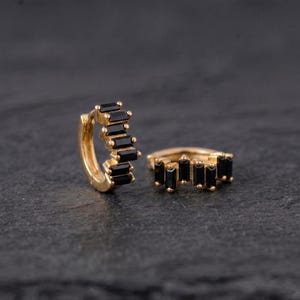 May include: A pair of gold hoop earrings featuring a row of rectangular black baguette-cut stones set along the outer edge. The earrings have a hinged closure mechanism and a polished metallic finish, showcasing a series of dark stones.