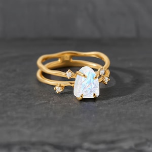 May include: A gold-colored ring set featuring a large, iridescent, triangular-shaped gemstone. The ring is accented with small, clear gemstones and has a double-band design. The ring is displayed on a dark gray surface.