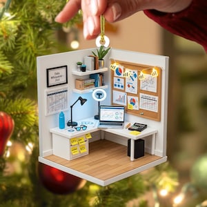 Pode incluir: Um enfeite de Natal de escritório em miniatura. O enfeite apresenta uma mesa com um laptop, uma lâmpada e uma calculadora. Um quadro de cortiça exibe gráficos e diagramas. O enfeite é preso por um fio dourado.