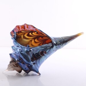May include: A handcrafted glass seashell sculpture. The shell features a swirling pattern of orange and yellow within, transitioning to a deep blue and purple exterior. The sculpture is a decorative art piece.