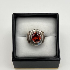 May include: A silver ring featuring a large, oval-cut, red gemstone. The ring is set in a silver band with a distinctive design, displayed in a black jewellery box with a white interior. The red gemstone is the central feature.