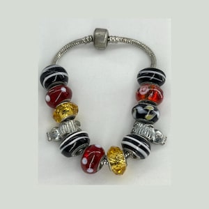 May include: A silver charm bracelet with a variety of colorful beads. The beads include black and white striped, red with white polka dots, and gold-colored beads. The bracelet is a unique piece of jewelry.