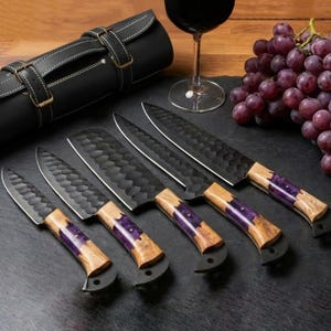 May include: A set of four kitchen knives with black hammered metal blades and handles crafted from light brown wood with a purple resin inlay. The knives are arranged in a row next to a black leather roll, a wine glass, and a bunch of red grapes.