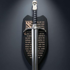 以下が含まれることがあります： 銀色の刃と黒い柄の装飾的な剣。柄頭には白く詳細なライオンの頭が描かれています。剣は、刻まれたテキストが書かれた黒い盾に取り付けられています。盾には装飾的なイニシャルがあります。