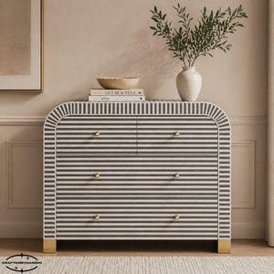 Puede incluir: Cómoda de cuatro cajones con un patrón de rayas blancas y negras. La cómoda tiene herrajes y patas doradas. Un jarrón con vegetación, un cuenco y libros están encima.