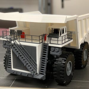 Op de afbeelding: Gedetailleerd model van een grote witte en grijze dumper. De vrachtwagen heeft een cabine, een grote laadbak en oversized zwarte banden. Het heeft een ladder en relingen voor toegang. Het model bevat rode details en staat op een donkere ondergrond.