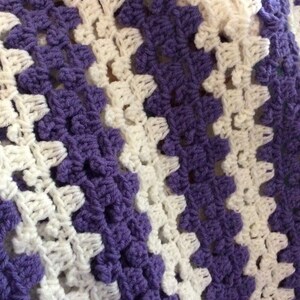 May include: A crocheted blanket with alternating stripes of white and purple yarn. The blanket is made with a simple stitch pattern and has a soft, textured feel.